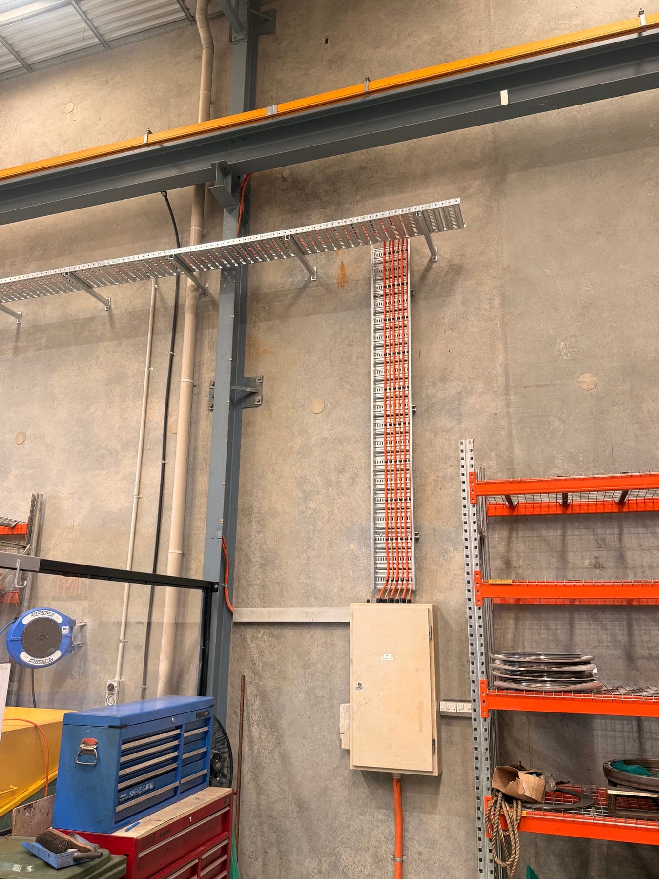 A galvanised cable tray carrying a bank of orange conduits down to a wall-mounted sub-board in a warehouse.