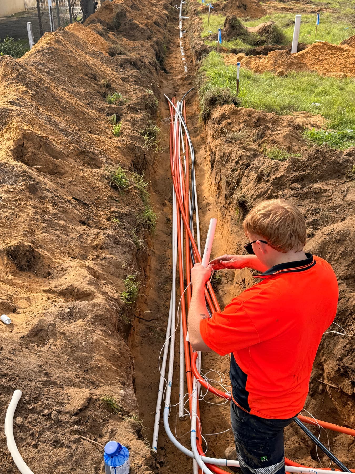 A Brady electrician in hi-vis laying orange electrical conduit along a sand service trench on a new-build site.